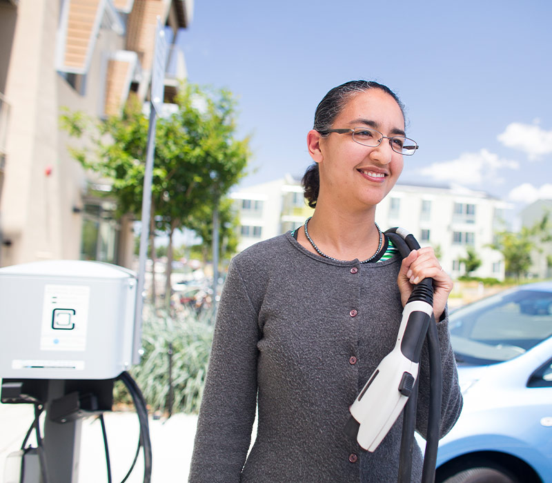 A woman holds an electric car charging cable as she walks towards her car.